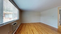 This 2-bedroom, 1-bathroom single-floor unit offers a generous and well-distributed layout across its living spaces. Warm-toned hardwood flooring runs throughout the main living areas and bedrooms, contributing to a cohesive and inviting aesthetic. Neutral-painted walls provide a clean, versatile backdrop throughout.

The living room is notably spacious, featuring large windows fitted with blinds that allow abundant natural light to fill the room. A wall-mounted HVAC unit and baseboard heating provide climate control. The living area connects openly to the kitchen, which is equipped with white cabinetry, dark countertops, a double-basin sink, and white appliances including a refrigerator, electric range, built-in microwave, and dishwasher. A window above the sink adds natural light to the cooking space, and tile flooring grounds the kitchen area.

Both bedrooms continue the hardwood flooring theme and are well-sized. One bedroom features a set of louvered bifold closet doors, offering practical storage. Each room includes windows with blinds and baseboard heating. The bathroom includes a full bathtub with white tile surround and a towel bar, maintaining a clean, functional design throughout.