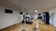 This dedicated fitness space offers a comprehensive workout environment housed within a single open-plan floor. The room is defined by its polished, stained concrete flooring, which features a warm amber tone with natural variation throughout — a durable and low-maintenance surface well-suited for a high-activity setting. Structural support columns, painted in a variety of colors including teal, green, and orange, punctuate the space and add a subtle visual character to the otherwise clean, white-walled interior.

The cardio section is well-appointed, featuring treadmills, a stationary bike, and a rowing machine, providing a range of aerobic training options. A dedicated strength training area includes a multi-station cable machine, a weight bench, and a tiered dumbbell rack stocked with a full range of free weights. Additional equipment such as a pull-up/dip station rounds out the strength offerings.

Multiple flat-screen televisions are mounted along the walls, ensuring visibility from virtually any point in the room. Recessed and panel lighting illuminates the space evenly from above, while a small louvered window on one wall provides a source of natural ventilation. The layout allows for generous open floor space between equipment stations, supporting a comfortable and functional flow throughout the facility.