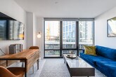 This well-designed single-floor unit showcases a cohesive modern aesthetic defined by crisp white walls, light wood-toned flooring, and thoughtful detailing throughout. The bedroom serves as a comfortable retreat, featuring a dark-upholstered bed frame paired with layered neutral bedding and accent pillows. A bold blue abstract artwork anchors the wall above the headboard, while modular black metal wall shelves add functional decor on the opposite side. A globe bedside lamp provides warm ambient lighting, and a mirrored closet is accessible through a clean white door. The bathroom is finished in a minimalist style, featuring light tile flooring, a glass-framed shower enclosure with dark metal hardware, a wall-mounted toilet, and a dark wood vanity with a stone-look countertop. The kitchen is equipped with stainless steel appliances, including a full-size refrigerator, built-in oven, microwave, and induction cooktop. Walnut-toned lower cabinetry contrasts elegantly with white upper cabinets and a light stone backsplash. The living area features a rich navy velvet sofa accented with gold and teal cushions, complemented by abstract artwork and decorative wall shelving. Floor-to-ceiling windows allow natural light to pour in from an adjacent outdoor area.