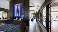 This single-floor amenity space makes a striking first impression with a sleek reception area anchored by a bold blue "M" logo artwork, dark wood paneling, and rich hardwood flooring that extends throughout. The entryway flows naturally into an expansive lounge and social hub, where soaring vaulted ceilings with exposed beams draw the eye upward. The lounge is boldly designed with vibrant cobalt blue carpeting, curved sectional sofas, sculptural accent chairs, and a statement chandelier, all complemented by oversized tropical plants that add a lush, organic touch. A built-in workstation area with teal seating offers a dedicated space for business or focus. Adjacent to the lounge, a well-appointed kitchenette features marble-style countertops, a Starbucks coffee machine, under-counter refrigeration, and a geometric-patterned accent wall that adds artistic depth. The fitness center is generously sized and filled with natural light from large windows, showcasing vaulted ceilings with ceiling fans, white painted brick walls, and a striking mural ceiling detail. It is equipped with a comprehensive array of cardio machines and strength training equipment on durable red and gray rubber flooring. A utilitarian corridor with white brick walls, red rubber flooring, and dual water fountains provides convenient access to restrooms and exits.