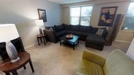 This single-floor space offers a warm and functional living environment designed for everyday comfort. The living area serves as the centerpiece of the home, featuring plush wall-to-wall carpeting that adds a sense of coziness underfoot. A large window allows natural light to flow into the room, brightening the neutral-toned walls and creating an airy atmosphere. The space is anchored by a generously sized dark sectional sofa, providing abundant seating and a natural focal point for relaxation or entertaining. A matching dark coffee table sits at the center, complemented by a coordinating accent chair in an olive tone that adds a subtle contrast to the room's palette. Decorative wall art adorns multiple walls, contributing character and visual interest to the space. A classic table lamp on a round side table offers warm ambient lighting for evening hours. The overall ambiance is casual yet thoughtfully arranged, balancing comfort and style in a cohesive manner. The open feel of the living area, combined with its natural light and soft furnishings, makes this a welcoming and practical space suited for a variety of lifestyles.