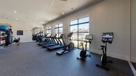 This single-level fitness center is thoughtfully designed to accommodate a comprehensive range of workout activities. The main exercise floor features durable speckled rubber flooring throughout and is organized into distinct functional zones. Large black-framed windows line the walls, flooding the space with natural light and offering views of the surrounding neighborhood, while ceiling fans provide additional ventilation and comfort.

The cardio zone is well-appointed with multiple treadmills, elliptical machines, and stationary cycling bikes, all positioned to face the oversized windows. The strength training area includes a full rack of dumbbells, multiple selectorized machines, a multi-functional cable system, and weight benches. A climbing wall is integrated into one section, adding a dynamic functional fitness element. Resistance training accessories, medicine balls, and additional equipment are neatly organized on dedicated storage racks.

A separate amenity area features teal cabinetry with open shelving for storage, a beverage refrigerator, a utility sink, and a water fountain station. A mirrored section enhances the sense of space. Recessed lighting and ceiling fans complement the natural light throughout. An additional room, accessible via a teal door, provides supplementary space for varied fitness activities.