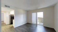 This single-floor space features a clean, minimalist design with a focus on functionality and natural light. The main living area is finished with dark hardwood-style flooring that contrasts attractively against the neutral white walls, creating a contemporary yet understated aesthetic. A textured popcorn ceiling adds a classic touch overhead.

A sliding glass door fitted with vertical blinds serves as a prominent architectural feature, allowing natural light to flood the interior while providing direct access to a private outdoor balcony. The balcony, enclosed by a railing, offers an outdoor extension of the living space. The vertical blinds provide flexible light control and privacy as needed.

The open layout of the living area allows for versatile furniture arrangement and a spacious feel. A partial view toward the left side of the space suggests an adjacent kitchen or utility area, hinting at a practical, connected floor plan. The overall ambiance is bright and neutral, presenting a move-in ready environment with a simple, clean palette suitable for a variety of interior design styles.