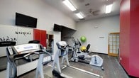 This fitness center offers a practical and motivating environment for residents or members looking to maintain an active lifestyle. The space is designed with functionality in mind, featuring a selection of cardio equipment including treadmills and elliptical machines arranged to maximize floor space. A ceiling fan and overhead fluorescent lighting ensure the room remains comfortable and well-lit during workouts.

The walls are kept clean and neutral, with a bold "motivate" wall decal serving as an inspirational focal point. A large wall mirror on one side visually expands the space and allows users to monitor their form during exercise. Two wall-mounted flat-screen televisions are positioned for easy viewing from the cardio equipment, adding an entertainment element to workouts.

Storage-friendly touches include stability balls in green and yellow tucked neatly in the background. The flooring features durable, multi-toned carpet tiles suited for a high-traffic fitness environment. A red accent door adds a pop of color to the otherwise neutral palette. Overall, the space strikes a balance between compact efficiency and a welcoming, energizing atmosphere.