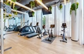 Located in the vibrant Wynwood district of Miami, this thoughtfully designed fitness center offers a well-appointed workout environment that seamlessly blends function with aesthetic appeal. The space features light wood-plank flooring throughout, providing a warm contrast to the dark, exposed ceiling that houses industrial-style ductwork and sleek linear lighting fixtures.

A standout design element is the bold, abstract mural adorning one wall, featuring soft earth tones of blush, terracotta, and grey — adding an artistic, gallery-worthy touch befitting the Wynwood neighborhood. Lush hanging greenery and moss ball installations suspended from the ceiling introduce a biophilic element that breathes life into the interior.

Floor-to-ceiling sheer curtains line the exterior wall, filtering abundant natural light and framing large glass doors that open directly onto a manicured outdoor green space with lounge seating — blurring the boundary between indoors and out. The gym is fully equipped with treadmills, stair climbers, stationary and spin bikes, a cable machine, a power rack, and a full dumbbell rack. Mounted flat-screen TVs provide entertainment throughout. The overall ambiance is bright, modern, and energizing — a premium wellness experience in the heart of Miami.