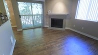 This single-floor living space offers a clean, well-appointed interior centered around a standout fireplace framed in light grey tile with a crisp white mantle, serving as the room's focal point. The living area is finished with warm-toned hardwood flooring that runs throughout, adding both character and a sense of continuity to the space. Neutral-toned walls complement the natural materials, creating a balanced and inviting atmosphere. A large sliding glass door floods the room with natural light and provides direct access to a balcony, beyond which mature trees and greenery create a private, serene outdoor backdrop. Sheer curtains frame the door, softening the transition between indoors and out. White baseboards provide a clean contrast against the warm flooring and taupe walls. The layout of the living area is open and unobstructed, allowing for flexible furniture arrangements. Overall, the space combines practical finishes with comfortable design elements, making it a functional and aesthetically cohesive living environment suited for everyday living.