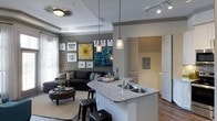 This single-floor space showcases a well-designed open-concept layout that seamlessly blends the kitchen and living areas into one cohesive environment. The kitchen features a striking granite-topped island with a built-in stainless steel sink, offering both functionality and style. Sleek pendant lights hang above the island, adding a modern touch, while track lighting illuminates the adjacent area. Crisp white cabinetry and clean lines contribute to the kitchen's polished aesthetic.

The living area flows naturally from the kitchen, anchored by a deep charcoal sectional sofa paired with a tufted leather ottoman, creating a comfortable and inviting atmosphere. A curated gallery wall displays a mix of framed artwork and bold canvases, adding personality and visual depth to the space. An ornate starburst ceiling fixture draws the eye upward to the tray ceiling detail, which adds architectural character throughout.

Warm hardwood flooring runs through the space, complementing the neutral gray walls. Natural light filters in through window treatments, brightening the interior. A hallway visible from the main area leads to additional rooms, suggesting a well-organized floor plan. Overall, this space balances contemporary design with comfortable livability, offering a refined yet approachable atmosphere.