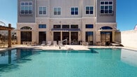 This amenity space combines an outdoor swimming pool with a well-appointed fitness center, delivering a comprehensive recreational experience. The sparkling pool features crystal-clear turquoise water and is framed by a spacious concrete deck lined with lounge chairs, providing ample room for relaxation. An ADA-accessible entry ramp ensures inclusive access. The pool is set against the backdrop of a multi-story building clad in brick and gray siding, with large windows and navy awnings that lend a polished, modern aesthetic to the outdoor environment.

The fitness center is thoughtfully divided into distinct workout zones. A dedicated cardio area features treadmills positioned beneath a wall-mounted television, surrounded by large windows that admit generous natural light and outdoor views. The strength training area is generously equipped with a power rack, cable machine, Precor stationary bike, rowing ergometer, dumbbells, kettlebells, medicine balls, battle ropes, and resistance bands. Full-length mirrors amplify the sense of space and allow for form checks during exercise. Ceiling fans maintain airflow throughout, and patterned herringbone carpet tiles add a contemporary design touch underfoot. The overall ambiance is bright, functional, and well-maintained.