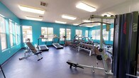 This fitness center offers a comprehensive workout environment designed to accommodate a wide range of exercise needs. The space is defined by striking teal accent walls that energize the atmosphere, complemented by a neutral grey rubber flooring that provides both durability and comfort underfoot. Bright fluorescent lighting ensures excellent visibility throughout the room, and multiple windows along the exterior wall allow natural light to fill the space while offering views of the surrounding outdoor greenery.

The facility is well-stocked with a variety of equipment. Cardio options include treadmills and an upright stationary bike, each positioned to take advantage of the natural light from the windows. A full dumbbell rack lines one wall, offering an extensive range of free weights alongside kettlebells, catering to both beginner and advanced users. An adjustable weight bench and a dedicated flat bench provide versatile options for strength training. A multi-functional cable and pull-up station rounds out the equipment selection, supporting a broad array of resistance exercises.

A large wall mirror spans a significant portion of the room, enhancing the sense of space and allowing users to monitor their form during workouts. The layout is thoughtfully organized to maximize floor space and ensure safe movement between stations, making this fitness center a practical and motivating environment for daily exercise.