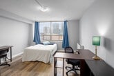 Located on the 4th floor at 186 King Street in the heart of downtown London, Ontario, this studio unit offers a practical and efficient open-concept layout designed to maximize every square foot. Light-toned luxury vinyl plank flooring runs throughout, lending a clean, cohesive look to the entire space.

The main living area serves multiple functions, combining sleeping, studying, and living zones in a single open room. A large window dressed with blue curtains fills the space with natural light while framing impressive views of the downtown London skyline. A dedicated workspace with a desk, ergonomic chair, and open shelving unit provides a functional study area.

The kitchenette is neatly integrated into the space and features white cabinetry, a gas range, refrigerator, and a sink — offering all essential cooking amenities in a compact footprint. The layout ensures the kitchen does not intrude on the living or sleeping areas.

The private bathroom is well-appointed with wood-look flooring, a single vanity with a marble-style countertop, a toilet, and a shower with a curtain enclosure, maintaining a clean and neutral aesthetic throughout.

The unit's crisp white walls, neutral palette, and efficient use of space make it a functional and comfortable residence in a prime downtown location.