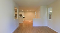 This single-floor space offers a clean, open-concept layout designed for effortless flow between living and kitchen areas. The interior is finished with warm light hardwood flooring that extends throughout, creating a cohesive and inviting atmosphere. Recessed lighting is installed across the ceiling, providing even, well-distributed illumination throughout the main living space.

The kitchen is thoughtfully appointed with white cabinetry that reaches toward the ceiling, dark countertops, and stainless steel appliances including a refrigerator and microwave. A kitchen island or peninsula adds additional prep and seating space, further connecting the kitchen to the open living area. A pass-through window cutout in the wall enhances the sense of openness between rooms.

The living area is generously proportioned, with smooth white walls and clean architectural lines that lend the space a modern, minimalist character. A small window near the rear of the space brings in natural light, complementing the recessed fixtures above. White baseboards trim the perimeter, adding a crisp finishing touch. Overall, the space presents a bright, functional, and well-maintained interior well-suited for everyday living.