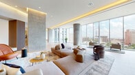 This single-floor amenity space is defined by its sweeping, open-plan layout and a refined modern aesthetic that balances scale with warmth. Soaring floor-to-ceiling glass walls wrap around multiple lounge areas, flooding the interiors with natural light and framing panoramic cityscape views. Polished concrete cylindrical columns punctuate the open spaces, contrasting beautifully against light wood-plank flooring and warm cove lighting integrated into tiered ceiling details throughout.

The main lounge area offers generous sectional seating arrangements on textured area rugs, complemented by sculptural accent chairs and gold-toned coffee tables. Adjacent to this, a more casual dining nook features long communal tables with sleek chairs positioned alongside the glass perimeter. A dedicated games room includes a billiards table set beneath curved ceilings with warm recessed lighting and wood-paneled walls, alongside a kitchenette with built-in appliances.

Transition corridors are thoughtfully designed with vertical wood slat screens, curated artwork, and intimate seating alcoves with tufted banquettes in teal upholstery. Corner window areas with large-format tile flooring provide sun-drenched retreats with unobstructed urban views. The overall ambiance is polished, airy, and inviting.