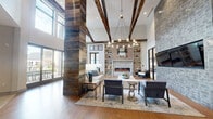 This thoughtfully designed single-floor communal space masterfully blends rustic character with contemporary sophistication. Soaring double-height ceilings draw the eye upward to dramatic exposed wood beam trusses that stretch across the open volume, creating a striking architectural focal point. Reclaimed wood-clad columns anchor the layout, adding warmth and texture that contrasts beautifully with the crisp white walls and modern finishes throughout.

The main lounge area centers around a built-in fireplace framed by a detailed brick surround and flanked by open shelving, with a large-format television mounted on an adjacent grey tile accent wall. A circular chandelier with warm-toned glass pendants illuminates the seating arrangement below, which includes a neutral sofa, upholstered accent chairs, and a central coffee table set over a layered area rug on wide-plank hardwood flooring.

A secondary zone transitions to large-format tile flooring and features a shuffleboard table alongside a wall-mounted display screen set against grey paneled wainscoting, offering a dedicated recreation area. Floor-to-ceiling black-framed windows and sliding glass doors flood the entire space with abundant natural light while providing views of an outdoor terrace and surrounding greenery, seamlessly connecting the interior to the exterior environment.