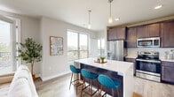 This single-floor residence showcases a thoughtfully designed open-concept layout with contemporary finishes throughout. The kitchen serves as a central highlight, featuring rich dark-toned cabinetry, white quartz countertops, a full suite of stainless steel appliances including a French door refrigerator, gas range, microwave, and dishwasher, as well as a subway tile backsplash. A spacious island with additional storage provides casual seating for three, illuminated by sleek pendant lighting. Large windows adjacent to the kitchen flood the space with natural light and offer elevated outdoor views of surrounding greenery.

The living area is bright and welcoming, with light wood-style flooring, a ceiling fan, and direct access to a private balcony through full-length glass doors fitted with horizontal blinds. The bedroom features plush carpeting, neutral walls, and generous space, with a ceiling fan and direct access to a well-finished bathroom. The bathroom includes a wide vanity with quartz countertops, dark cabinetry, a large mirror with pendant lighting, wood-look tile flooring, and a shower-tub combination. Throughout, the space maintains a clean, modern aesthetic with a neutral palette and quality finishes.