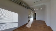 This single-floor space showcases a striking industrial-modern aesthetic that blends raw architectural elements with clean, contemporary design. The open-concept layout creates a generous, airy feel, unified by rich hardwood flooring that runs throughout and catches natural light pouring in through large windows fitted with horizontal blinds.

Soaring ceilings draw the eye upward to a prominent exposed metal ductwork system that runs the length of the space, serving as both a functional and decorative feature. A cluster of industrial-style pendant lights hang at varying heights, providing warm, focused illumination and reinforcing the space's urban character.

The kitchen area is thoughtfully integrated into the open floor plan, featuring white cabinetry, a gas range, and under-cabinet ventilation — offering a clean, functional workspace without interrupting the flow of the broader living area. The crisp white walls throughout provide a neutral backdrop that amplifies the sense of light and openness.

A hallway visible toward the rear of the space suggests additional rooms or utility areas, adding to the overall functionality of the layout. The combination of natural light, warm wood tones, and exposed architectural details gives this space a distinctive, loft-inspired character that balances industrial edge with livable warmth.