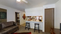 This single-floor residence showcases an open-concept design that connects the living, dining, and kitchen areas into one cohesive space. The kitchen is positioned behind a raised white pass-through counter, which doubles as a breakfast bar accommodating two black bar stools — an efficient and social setup ideal for casual dining or entertaining. The kitchen itself is equipped with a cooktop range, refrigerator, and cabinetry, with a mirrored backsplash that adds depth and reflects light throughout the space. The counter surface provides functional workspace for meal preparation.

The living area flows naturally from the kitchen bar, featuring neutral-toned carpet underfoot and white walls that keep the atmosphere bright and clean. A textured popcorn ceiling runs throughout, characteristic of the building's construction era. Colorful wall art adds a decorative focal point to the shared space.

A doorway visible from the main living area leads directly into the bedroom, suggesting a practical, straightforward floor plan. A wood-toned door near the kitchen area likely accesses a closet or utility space. Overall, the layout is compact yet functional, maximizing the use of available space across a single level.