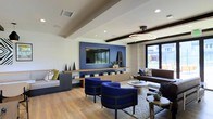 This single-floor space presents a thoughtfully designed modern lounge interior that balances bold design choices with comfortable functionality. The room is anchored by a striking blue accent wall, which frames a large flat-screen television mounted above a wooden media console. The console provides ample display and storage space, decorated with curated décor items that add personality to the setting.

Light wood flooring runs throughout, lending warmth and continuity to the space. The ceiling features exposed dark wood beams and a tray detail, adding architectural depth, while recessed lighting and a contemporary geometric pendant fixture ensure the room is well-lit throughout. A large window on one side invites natural light and offers views of the surrounding exterior.

Seating is varied and inviting, with a pair of cobalt blue accent chairs positioned centrally on a textured area rug, complemented by a dark brown leather sofa and a built-in bench seating area. Round side tables and a low circular coffee table complete the arrangement. The overall ambiance is contemporary and vibrant, suited for communal relaxation or informal gatherings.