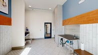 Located at 1300 Front Ave in Columbus, Georgia, this dedicated pet grooming room is thoughtfully designed to accommodate all aspects of pet care in a clean and functional environment. The space features soaring ceilings that lend an open, airy feel despite its compact footprint, with recessed lighting providing bright, even illumination throughout.

The interior is defined by a striking two-tone tile treatment: vertical white subway tiles line the lower walls, transitioning to a bold band of orange ceramic tiles at mid-height, set against cool blue-gray upper walls. The white grid-pattern tile flooring is both practical and easy to maintain, complementing the overall aesthetic.

Two primary workstations anchor the room — a stainless steel grooming table equipped with a built-in dryer and grooming accessories, and a deep stainless steel wash basin with a step stool for easy pet access. A stainless steel trash receptacle and wall-mounted towel bars add to the room's functionality. Framed animal-themed artwork decorates the walls, adding a welcoming touch.

Natural light filters in through large windows and a glass-panel exterior door, connecting the space to the outdoors. A secondary interior glass door provides access to the adjacent building. The overall ambiance is bright, hygienic, and professionally appointed, making this an ideal self-serve or full-service pet grooming facility.