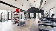 Located at 651 W 42nd Ave in Denver, CO, this Wellness Club offers a generously sized, single-floor exercise facility designed to accommodate a wide range of fitness activities. The space is defined by its striking vaulted ceilings, which rise dramatically overhead and are accented by copper pendant lights and ceiling fans, lending an industrial-chic aesthetic complemented by dark corrugated metal wall panels.

Large, black-framed windows and glass doors line the perimeter, flooding the interior with natural light and providing views of the surrounding outdoor areas. The flooring transitions between light wood and rich dark hardwood, delineating different workout zones throughout the space.

Equipment is comprehensive and thoughtfully arranged. A central functional training rig supports TRX suspension trainers, a hanging heavy bag, and resistance attachments with yellow straps. Surrounding this hub are kettlebells, medicine balls, sandbags, battle ropes, and plyometric boxes for functional and strength training. A dedicated cardio zone features treadmills, stationary bikes, rowing machines, and an elliptical, while a strength area includes Matrix selectorized machines and a full rack of dumbbells. The facility also incorporates a tire-flip training tool and a separate, quieter cardio room with motivational wall art, rounding out this versatile, full-service fitness environment.