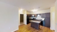 This single-floor residence offers a thoughtfully designed layout centered around an open-concept kitchen that serves as the heart of the space. The kitchen is outfitted with rich, dark wood cabinetry that extends to the upper walls, including a distinctive built-in wine rack that adds a refined touch. Granite countertops provide generous prep space, while the central kitchen island offers additional workspace and casual seating potential. Black appliances — including a refrigerator, range, microwave, and dishwasher — lend a sleek, cohesive aesthetic throughout the kitchen. Elegant pendant lighting hangs above the island, complementing the overhead flush-mount fixtures and creating a warm, inviting atmosphere. The warm-toned hardwood flooring flows seamlessly through the main living area, enhancing the sense of openness and continuity. A doorway visible from the kitchen leads into an adjoining carpeted room, suggesting a comfortable and well-connected floor plan. The smooth, light-colored walls and neutral palette throughout contribute to a bright and airy ambiance, making the space feel welcoming and versatile. Overall, this residence combines functional design with tasteful finishes for comfortable everyday living.