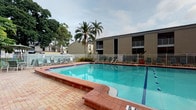 This community amenity space offers residents two distinct areas designed for active, healthy living. The outdoor pool area is a standout feature, showcasing a generously sized swimming pool with clearly marked depth indicators and a classic brick-paved surround. Lounge chairs are arranged along the pool deck, providing ample space for relaxation. Tall palm trees and mature landscaping frame the area, creating a tropical ambiance, while a white perimeter fence ensures privacy and safety.

The fitness center is accessed through a secured, keypad-entry door and is well-organized for a comprehensive workout experience. The space is equipped with cardio machines including a treadmill and an elliptical trainer, as well as a multi-station cable weight machine with an adjustable bench. A large wall-mounted mirror enhances the sense of space and allows for form-checking during exercise. A dedicated functional training rack holds an extensive collection of accessories, including medicine balls, resistance bands, stability balls of varying sizes, and foam rollers. A wall-mounted flat-screen television adds convenience for entertainment during workouts. The flooring throughout is durable rubber gym matting, and natural light filters in through windows, keeping the atmosphere bright and motivating.