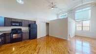 This single-floor unit showcases an open-concept layout that maximizes the sense of space, anchored by warm, honey-toned hardwood floors that run throughout the main living areas. Soaring ceilings lend an airy, loft-like quality to the space, accentuated by arched transom windows that invite natural light deep into the interior. A ceiling fan with a light fixture serves the main living area, enhancing both comfort and airflow.

The kitchen is well-appointed with rich, dark espresso-toned cabinetry featuring sleek hardware, granite-look countertops, and a full suite of black appliances including a refrigerator, range, dishwasher, and microwave. A stainless steel sink with modern faucet completes the functional workspace, while ample upper and lower cabinet storage ensures excellent organization.

The open living area provides generous room for flexible furniture arrangements, with a baseboard heating unit along one wall offering efficient climate control. A built-in closet with a rod offers convenient storage within the main living space.

The bathroom is clean and straightforward, featuring a tub-shower combination, a white vanity with sink, a mirrored medicine cabinet, and warm neutral wall tones. Durable tile-look flooring completes the space with easy maintenance in mind. Overall, this unit combines practical amenities with distinctive architectural character.