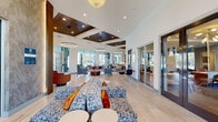 Located in Ocala, Florida, this single-story community clubhouse at 3585 SW 38th Terrace is a thoughtfully designed gathering space that blends contemporary aesthetics with functional amenities. The main lounge is an expansive, open-concept area anchored by light wood-look tile flooring, soaring ceilings, and a striking combination of white walls and rich wood-paneled ceiling accents. Oversized floor-to-ceiling windows and glass sliding doors flood the interior with abundant natural light while offering sweeping views of the resort-style pool and surrounding community. Stylish seating arrangements throughout the lounge feature patterned sofas, accent chairs in warm earth tones, and sculptural side tables, creating a welcoming social atmosphere. A dedicated kitchenette area is outfitted with modern cabinetry, a built-in coffee station, a mosaic tile backsplash, and a wall-mounted television — ideal for casual gatherings. A separate conference room provides a more intimate setting, complete with a round table, leather seating, patterned wallcovering, a Sputnik-style chandelier, and a wall-mounted display screen. Large decorative pendant lighting throughout adds a refined, boutique-hotel quality to the overall ambiance. The space seamlessly connects indoor comfort with outdoor living, making it an inviting hub for residents.