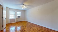 This single-floor residence showcases classic parquet hardwood flooring that runs throughout the space, finished to a high gloss that reflects natural light and adds warmth to every room. The generously sized main living area is bright and open, with neutral walls and white trim that create a clean, airy atmosphere. A ceiling fan with integrated light fixture provides both comfort and illumination, while windows fitted with window air conditioning units ensure year-round climate control. A radiator along the wall provides additional heating. An adjacent room, which can serve as a bedroom, continues the parquet flooring and features a window unit, a ceiling fan, and access to a spacious built-in closet with shelving — offering ample storage. The kitchen is compact yet functional, equipped with white cabinetry, a gas range, and a refrigerator, with subway tile backsplash detailing. The bathroom is finished in classic white subway tile with black accent trim, and includes a full soaking tub with shower, a pedestal sink, white floor tile, and a window for natural ventilation. Clean lines and timeless finishes define the overall aesthetic of this well-maintained single-floor residence.