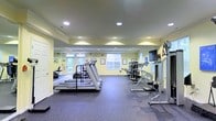This single-floor fitness center offers a comprehensive workout environment designed to accommodate a variety of exercise routines. The space is finished with soft yellow walls and durable rubber flooring, creating a clean and functional atmosphere suited for physical activity. Recessed ceiling lights provide consistent, even illumination throughout the room, complemented by natural light streaming in through multiple large windows that lend an open, airy feel to the interior. The cardio area is anchored by a row of treadmills positioned toward the center of the space, offering ample room for users to exercise comfortably. Toward the rear and sides, a selection of strength and resistance training machines are arranged to allow for efficient use of the floor plan. A wall-mounted flat-screen television is positioned for visibility during workouts. Framed artwork decorates the walls, adding a welcoming touch to the otherwise utilitarian environment. The layout is thoughtfully organized to provide clear sightlines across the room, ensuring ease of movement between equipment. Overall, the fitness center presents a practical and inviting space that caters to a range of fitness needs within a well-maintained, single-level setting.