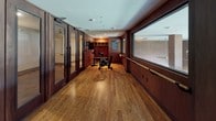 This fitness studio is designed with both functionality and aesthetic refinement in mind. The space is finished with rich, warm-toned hardwood flooring throughout, complemented by dark wood wall paneling that lends the room a sophisticated, boutique-gym atmosphere. Large framed windows line one side of the elongated space, allowing natural light to filter in and offering views into an adjacent room, enhancing the open and connected feel of the facility.

The layout is organized along a long, narrow floor plan that accommodates a variety of workout modalities. A mounted ballet barre runs along the windowed wall, supporting dance-style fitness and stretching activities. Cardio equipment, including stationary cycling bikes, is positioned near the barre section. Along the opposite side, a well-stocked free weights area features a tiered dumbbell rack alongside kettlebells, medicine balls, and resistance accessories neatly arranged on storage units. Built-in cubbies with hooks provide convenient storage for personal belongings.

Tall, glass-paneled double doors with dark wood framing serve as the main entry, adding a polished architectural detail. Recessed lighting overhead ensures consistent illumination throughout, while informational signage is displayed on the walls. The overall ambiance is clean, professional, and thoughtfully organized.