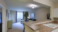 This single-floor residence showcases an open-concept layout that seamlessly connects the kitchen and living areas, creating a cohesive and functional environment. The kitchen features a broad peninsula with a granite-patterned countertop, offering both additional workspace and a natural partition between the kitchen and the adjoining living space. A built-in sink is integrated into the peninsula, adding practical utility to the design.

The living area is carpeted in a neutral tone, contributing to a warm and comfortable ambiance. A large window dressed with blue-grey curtains allows natural light to fill the room, brightening the soft grey walls throughout. The space is furnished with a mix of seating options, including a patterned accent chair and a dark sofa, complemented by green accent chairs that add a subtle pop of color.

A tall indoor plant near the window adds a natural, organic element to the interior. Framed wall art and a classic floor lamp contribute to a well-appointed, livable atmosphere. Ceiling-mounted flush lighting provides consistent illumination, ensuring the space remains bright and inviting throughout the day.