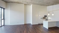 This single-floor space showcases a compelling blend of industrial and contemporary design elements. The exposed concrete ceiling serves as a striking architectural focal point, lending the space a raw, urban character that is balanced beautifully by the warmth of the rich, dark hardwood flooring throughout. Crisp white walls amplify the sense of openness and allow natural light to reflect generously across the interior.

The open-concept layout seamlessly connects the living area to the kitchen, creating a fluid and functional living environment. The modern kitchen is outfitted with sleek white cabinetry that extends to the ceiling, offering ample storage. Stainless steel appliances, including a microwave and range, are integrated neatly into the cabinetry, while a clean countertop workspace and sink complete the functional yet stylish setup. A kitchen island or peninsula adds an additional surface ideal for casual dining or meal preparation.

The generous open living area provides a versatile canvas, well-suited for a variety of furniture arrangements. The interplay between the industrial concrete ceiling and the refined finishes below gives the space a distinctive, design-forward character that is both modern and inviting.