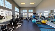 Located at 807 Central Park Circle in Kalamazoo, Michigan, this single-story fitness center is designed to support a comprehensive range of workout routines. The space is defined by its dramatically tall ceilings and an abundance of natural light, drawn in through large multi-pane windows that line the upper and lower portions of the walls, offering views of the surrounding grounds. The gray-painted walls and dark rubber flooring create a clean, functional aesthetic throughout.

The cardio area is well-stocked with multiple treadmills, elliptical trainers, and a rowing machine, all positioned to take advantage of the natural light and outdoor views. A wall-mounted television provides entertainment during workouts. A dedicated strength training zone features a multi-station cable machine, while a full dumbbell rack with a wide range of weights serves the free weight area. Full-length mirrors span one wall, enhancing the sense of space and allowing users to monitor their form.

The open central floor area accommodates functional fitness accessories including stability balls, resistance bands, medicine balls, and exercise mats. The space is accessed through elegant French doors, adding a refined architectural touch. Overhead flush lighting supplements the natural light, ensuring the space remains bright and inviting at all times.