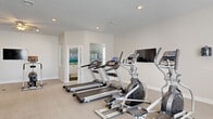 This single-floor fitness center is thoughtfully designed to support a comprehensive workout experience. The main exercise area is generously proportioned, featuring soft carpeted flooring throughout and bright, neutral walls that create a clean and energizing atmosphere. Recessed lighting and ceiling fans ensure the space remains well-lit and comfortable during any workout.

The cardio zone is outfitted with multiple treadmills and elliptical machines, all arranged to face wall-mounted flat-screen televisions, allowing users to stay entertained during their sessions. Adjacent to the cardio equipment, a dedicated strength training area houses a multi-station cable machine, a weight bench with a barbell rack, and a full dumbbell rack offering a range of weights. Protective rubber floor mats are positioned beneath the equipment for safety and durability.

Full-length mirrors line one wall of the strength area, enhancing the sense of space and allowing users to monitor their form. A ceiling fan overhead adds additional airflow to this zone.

A separate, connected room — distinguished by a vibrant mountain landscape mural — provides a welcoming retreat space or cool-down area, adding a creative and calming visual element to the facility. Natural light filters in through windows, contributing to the open, airy feel of the overall space.