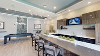 Located at 625 Spotswood Englishtown Rd in Monroe Township, NJ, this single-floor amenity center offers a thoughtfully designed collection of community spaces finished with contemporary style throughout. Light wood-look tile flooring and neutral tones set a clean, modern foundation across the facility.

The centerpiece is a spacious clubroom featuring a large granite-topped bar with seating for multiple guests, dark wood cabinetry, a white subway tile backsplash, pendant lighting, and a wall-mounted TV — ideal for social gatherings. Large windows with floor-to-ceiling drapes invite natural light and offer views of the surrounding greenery. A billiards table anchors one end of the room for recreation.

A dedicated lounge area provides a relaxed setting with a gray sectional sofa, accent chairs, and built-in shelving, complemented by curated wall art throughout.

The fitness center is generously proportioned and fully appointed with a wide array of strength-training machines, free weights, cable systems, and cardio equipment. Floor-to-ceiling mirrors, recessed lighting, and multiple wall-mounted TVs enhance the workout environment.

A business center nook offers dual computer workstations with ergonomic seating, set against a textured tile accent wall. The connecting hallways feature wainscoting, potted greenery, and geometric wallcovering accents, giving each transition area a polished, welcoming feel.
