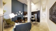 This contemporary single-floor residence showcases a thoughtfully designed open-concept layout that seamlessly integrates the living, dining, and kitchen areas into one cohesive space. Soaring ceilings amplify the sense of spaciousness, while polished concrete floors lend an industrial-chic aesthetic throughout.

The living area features a bold deep-teal accent wall adorned with decorative plate arrangements, creating a striking focal point. A sleek media console with a flat-screen television anchors the space, complemented by mid-century modern seating in soft blue-gray tones and a statement floor lamp that adds warmth.

The kitchen is outfitted with dark espresso cabinetry that extends to the ceiling, stainless steel appliances including a refrigerator, range, and microwave, and a clean countertop workspace. Structural white columns punctuate the open floor plan, adding architectural character without interrupting the flow between spaces.

A compact dining area with an industrial-style table and wire-frame chairs bridges the kitchen and living zones, creating a casual and functional setting. Natural light filters through the space, and a hallway visible in the background suggests additional private rooms beyond. The overall ambiance is modern, urban, and well-curated.