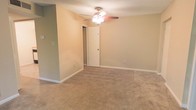 This single-floor space presents a straightforward and functional layout designed for comfortable everyday living. The main room is generously sized, featuring wall-to-wall carpeting in a warm beige tone that adds softness underfoot and contributes to the room's cozy atmosphere. Neutral-toned walls in a soft tan hue provide a versatile backdrop, making the space easy to personalize with a variety of décor styles. A ceiling fan with an integrated light fixture is centrally mounted, offering both practical airflow and ambient lighting throughout the room. The textured ceiling adds a subtle visual element characteristic of residential construction. White baseboards frame the perimeter of the room, providing a clean contrast against the warm wall color. A white interior door leads to an adjoining area or closet, hinting at additional storage or functional space beyond the main room. Electrical outlets are conveniently positioned along the walls. The overall ambiance of the space is calm and understated, offering a blank canvas that lends itself well to a range of uses, from a living area to a bedroom or multi-purpose room. The single-floor layout ensures easy accessibility and a cohesive flow throughout.