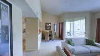 This well-appointed single-floor residence showcases an open-concept layout that connects the living, dining, and kitchen areas in a cohesive, airy design. The living room is anchored by a plush white sofa and a wicker accent chair, accented with terracotta and green decorative pillows that complement the green area rug and dark cube coffee table. Large sliding glass doors flood the space with natural light and open directly onto a private balcony, where outdoor seating provides views of lush palm trees beyond the railing. Rich, dark curtains frame both the balcony doors and the adjacent windows, adding a warm contrast to the neutral walls and soft carpeted floors. The soaring vaulted ceilings dramatically expand the sense of space throughout the main living area, creating an open and welcoming ambiance. The dining area is positioned adjacent to the living room and features a round table with wicker dining chairs beneath a pendant light, adding a casual yet refined touch. A half-wall partition defines the kitchen, which is visible with light cabinetry and a tiled backsplash. The overall aesthetic is a clean, contemporary style with warm accents, enhanced by abundant natural light and seamless indoor-outdoor connectivity.