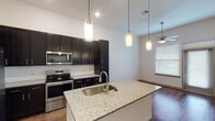 This well-appointed 1-bedroom, 1-bathroom unit in Houston, TX offers a contemporary living experience with thoughtful design details throughout. The open-concept kitchen serves as the centerpiece of the living area, showcasing rich dark cabinetry that extends to the ceiling for ample storage, granite countertops, and a full suite of stainless steel appliances including a French door refrigerator, electric range, and built-in microwave. A large kitchen island with a granite surface and undermount sink doubles as a breakfast bar, illuminated by a trio of modern cylindrical pendant lights. Warm hardwood-style flooring runs throughout the main living and kitchen areas, adding a sense of continuity and elegance. The bedroom is generously sized, featuring plush carpeting, a ceiling fan, and a large double-pane window with blinds that fills the room with natural light and offers an outdoor view. The bathroom is finished with granite countertops on the vanity, dark cabinetry, hardwood-style flooring, and a full soaking tub with tiled surround. A louvered door opens to a dedicated walk-in closet, providing convenient and organized storage. The unit's clean white walls and high ceilings enhance the open, airy ambiance throughout.