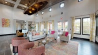 This expansive single-story communal space blends rustic character with modern design to create a welcoming and visually dynamic environment. The centerpiece of the main lounge is a dramatically high ceiling adorned with reclaimed wood beams and a suspended wooden canoe, adding a distinctive, conversation-worthy focal point overhead. Dark hardwood-style flooring flows throughout, grounding the space with warmth and continuity.

Multiple distinct seating zones cater to a variety of uses. One area features a light gray sofa and accent chairs accented with vibrant pink and patterned throw pillows, layered over a colorful area rug. Adjacent to this, a cluster of tan leather armchairs surrounds a round side table, creating a more intimate lounge setting. A reclaimed wood accent wall frames a mounted fireplace and television, serving as an architectural anchor for the space.

A dedicated dining area offers round tables paired with wooden chairs beneath pendant lighting, bathed in natural light streaming through large black-framed windows with views of lush outdoor greenery. Patterned yellow and white curtains frame the French doors that open to an outdoor terrace, further connecting the interior to its surroundings. Throughout the space, tall clerestory windows admit additional daylight, enhancing the open and airy atmosphere of this thoughtfully designed communal retreat.