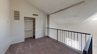 Located at 8028 S Central Ave in Phoenix, Arizona, this two-story unit offers a practical and efficiently organized layout spread evenly across two floors. The ground level hosts the main living and functional areas, anchored by a well-appointed kitchen featuring warm wood cabinetry, granite-style countertops, a double stainless steel sink, and black appliances including a refrigerator and range. A peninsula counter adds extra workspace and visual separation from the adjacent area. Stone-look tile flooring runs throughout the lower level, lending a durable and cohesive aesthetic. A staircase with a wrought iron railing connects the two floors, adding a structural focal point to the interior. The bathroom is cleanly finished with a dark wood vanity, white countertop, a large wall mirror, and a tub-shower combination. Upstairs, the upper-level room features plush carpeting, vaulted ceilings with a textured finish, and a built-in closet with open shelving for ample storage. A floor-to-ceiling sliding glass door floods the space with natural light and opens to an outdoor balcony area enclosed by a black metal railing, offering a connection to the outdoors. The overall design is straightforward and functional, well-suited for everyday living.