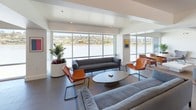 Located at 68 Barbaree Way in Belvedere Tiburon, California, this single-floor space offers a refined, contemporary design with a cohesive open layout spanning multiple functional zones. The interior is characterized by crisp white walls, recessed lighting, and large-format tile flooring throughout, creating a clean, professional aesthetic.

The main lounge area is a standout feature, showcasing expansive floor-to-ceiling windows that frame dramatic waterfront views of the surrounding bay and hillside landscape. This sun-filled seating area is furnished with plush gray sofas, cognac leather accent chairs, and a low oval coffee table, cultivating an inviting yet polished atmosphere.

Adjacent to the lounge, a sleek kitchen features a generous central island with a light stone countertop, white cabinetry, open wood shelving, and stainless steel appliances, providing ample functionality for catering or everyday use.

A dedicated conference and dining area accommodates a long solid-wood table surrounded by modern chairs, with glazed double doors offering direct access to the exterior. Smaller lounge alcoves provide additional seating for more intimate gatherings. The overall ambiance is modern, light-filled, and waterfront-inspired.