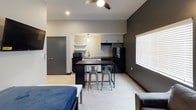 This single-floor studio unit showcases a contemporary design with a thoughtful open-concept layout that maximizes every square foot. The interior features a neutral color palette of light gray and white walls, accented by bold dark walls that add depth and modern contrast. Light wood-style flooring runs throughout the main living area, lending warmth to the space.

The kitchen is well-appointed with white cabinetry, dark countertops, and black appliances including a refrigerator, electric range with overhead ventilation, and a microwave. Open floating shelves provide additional storage and display space, while a stainless steel sink with a modern pull-down faucet completes the functional layout. A dark breakfast bar with industrial-style metal stools connects the kitchen to the living area, serving as both a dining space and a visual divider.

The main living and sleeping area is bright, benefiting from natural light through a sizable window. A ceiling fan with a light fixture enhances comfort, while a wall-mounted mini-split air conditioning unit ensures climate control year-round. A wall-mounted TV and a cozy seating area round out the living space.

The bathroom is clean and minimalist, finished with dark tile flooring and bright white walls. It includes a walk-in shower with grab bars, a floating vanity with a large mirror, and a toilet — designed with accessibility in mind.