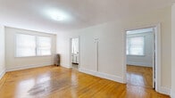 This single-floor unit offers a straightforward and functional layout across several well-defined rooms, unified by warm-toned hardwood flooring throughout the living areas and bedrooms. The main living space is generously proportioned, with crisp white walls, clean white baseboards, and multiple windows that invite natural light to fill the room. A classic cast-iron radiator provides character alongside the practical heating setup.

The kitchen is efficiently arranged, featuring white cabinetry, light countertops, a stainless steel sink, a gas range, and a tile backsplash in a neutral tone. A window above the range keeps the space bright and ventilated. The wood-look vinyl flooring in the kitchen offers a durable, easy-to-maintain surface.

The bedroom areas echo the same bright, neutral palette with hardwood floors, white walls, and windows fitted with blinds for privacy and light control. Each room benefits from natural light and offers a clean, move-in-ready canvas.

The bathroom is equipped with a full-sized soaking tub and overhead shower, a toilet, and a vanity with sink. Wood-look flooring and a bright window with blinds complete the space, keeping the overall aesthetic simple and functional throughout.