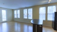 This single-floor space offers a clean, functional layout with a welcoming and versatile atmosphere. The main living area is generously sized, highlighted by a row of large double-hung windows along one wall that flood the interior with abundant natural light. The windows are fitted with horizontal blinds, allowing for adjustable light control while maintaining an airy feel throughout the space.

The walls are finished in a warm, neutral yellow-green tone that complements the light-toned hardwood-style flooring, which runs continuously across the space, adding warmth and a sense of cohesion. The smooth, light-colored ceiling features a visible vent, indicating a central HVAC system for year-round climate comfort.

A dark-finished, semi-circular counter or bar-height table is positioned near the window wall, providing a functional surface that contrasts elegantly against the lighter tones of the room. The open layout offers flexibility for furniture arrangement and multiple uses. White baseboards frame the perimeter neatly, adding a crisp, finished look. Overall, the space presents a bright, clean, and adaptable environment suited for a range of purposes.