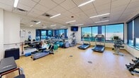 Located at 2700–2768 S Clermont Dr in Denver, Colorado, this single-floor fitness facility is thoughtfully designed to support a comprehensive workout experience. The space features a clean, modern aesthetic with drop-panel ceilings, recessed linear fluorescent lighting, and distinctive cork flooring throughout, providing both durability and a warm visual character.

The main workout area is generously sized, housing a variety of cardio equipment including multiple treadmills, an elliptical, a stationary bike, and a stair climber. A wall-mounted flat-screen TV keeps members entertained during workouts. Full-length mirrors line one wall, enhancing the sense of space. Large windows along the exterior wall admit abundant natural light and offer views of the surrounding Denver neighborhood, creating an open and energizing atmosphere.

Adjacent to the workout floor is a welcoming reception and multi-use area, featuring built-in light wood cabinetry, seating, and a wall-mounted television. Framed fitness and performance posters add a motivational touch to the décor. Glass-panel doors connect the areas, contributing to a transparent, open layout. Folding chairs and small workout accessories are readily accessible, indicating versatility for group or individual use. The facility is noted as open 24 hours.