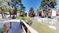 The outdoor common area at 2200 Lester Dr NE is a thoughtfully designed communal space that serves as a central gathering point within the property complex. The standout feature is a generously sized decorative pond, complete with multiple active fountains that create a calming water display and add a sense of vitality to the environment. Large natural boulders are strategically placed within the pond, contributing to the naturalistic aesthetic.

A well-constructed wooden walkway and bridge traverse the water feature, offering residents a pleasant path through the landscape. The walkway is fitted with sturdy railings, including glass panel sections that maintain open sightlines across the pond. Surrounding the water feature, carefully curated plantings include drought-tolerant desert plants, yucca, shrubs, and ground cover, creating a layered, low-maintenance landscape.

Mature pine and deciduous trees provide generous shade throughout the grounds, enhancing the outdoor comfort and contributing to a park-like atmosphere. Classic black lantern-style light posts are positioned along the pathways, ensuring the space is functional during evening hours. The multi-story residential buildings in warm red and white tones frame the perimeter, creating an enclosed, community-oriented setting that feels both private and inviting.