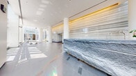 The lobby at 1188 Mission Street offers an impressive first impression through its sophisticated architectural design and high-end material palette. Soaring floor-to-ceiling glass curtain walls flood the space with abundant natural light, framing views of the surrounding San Francisco streetscape and creating an airy, open atmosphere. Tall, cylindrical white columns punctuate the interior, adding a sense of grandeur and structural elegance.

A dramatic feature wall clad in horizontally striated stone veneer, accented with warm gold trim, serves as a striking focal point and lends the space a refined, contemporary aesthetic. Complementing this is a substantial raw-edged granite reception fixture, whose natural, rough-hewn texture contrasts beautifully with the polished grey tile flooring below—a floor further distinguished by inlaid metallic accent tiles arranged in a geometric pattern.

Seating areas are furnished with sculptural marble benches and minimalist white stone stools, providing both function and artful detail. The lobby also includes access to a dedicated Amazon Hub Package Room, offering residents a convenient parcel retrieval service. Recessed ceiling lighting works in harmony with the natural light to maintain a bright, welcoming ambiance throughout. The overall design balances bold material choices with clean lines, resulting in a lobby that is both functional and architecturally compelling.