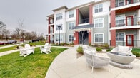 Located at 10951 Netherly Dr in Charlotte, NC, this residential community showcases a thoughtfully designed exterior that blends contemporary architectural elements with a welcoming neighborhood aesthetic. The multi-story buildings feature a striking combination of sage green siding, bold red accents, and white trim, creating a visually dynamic facade. Private balconies on upper levels offer residents elevated outdoor space with views of the surrounding grounds.

The community's outdoor areas are a standout feature, with smooth concrete pathways winding through lush, manicured green lawns and carefully maintained landscaping beds filled with shrubs, ornamental grasses, and seasonal plantings. Inviting seating areas are thoughtfully placed throughout, including classic white Adirondack chairs and modern woven lounge furniture positioned along the walkways for relaxed outdoor enjoyment.

A dedicated outdoor entertaining zone features built-in grilling stations and a ping pong table set on a spacious patio, ideal for social gatherings. A charming cottage-style leasing or amenity building with board-and-batten siding and large windows anchors one end of the property, complemented by low-profile bollard lighting along the paths. Ample surface parking is accessible and bordered by mature trees, providing a natural buffer. The overall ambiance is clean, modern, and community-oriented.