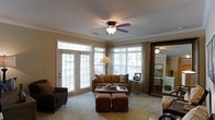 This single-floor space centers around a comfortable and thoughtfully designed living room that immediately draws the eye with its abundance of natural light. Large windows with white shutters line the walls, while a set of elegant French doors opens to the outdoors, flooding the room with warmth and providing a seamless connection to the exterior. Crown molding trims the ceiling throughout, lending a classic and polished architectural touch, while a ceiling fan with a built-in light fixture adds both style and practicality overhead. The living room is furnished with a warm-toned sofa accented by patterned throw pillows, a glass-top coffee table, and a pair of upholstered ottomans, creating an inviting and cohesive seating arrangement. A dark armchair anchors one corner of the room, complementing the rich, earthy color palette. Sheer curtain panels frame the windows, softening the natural light while maintaining an open and airy feel. Neutral wall tones and plush carpeting contribute to the overall warmth and comfort of the space. A large mirror near the entryway adds depth and reflects the natural light, enhancing the sense of spaciousness throughout.